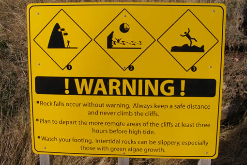 Yellow safety sign stating “! WARNING ! Rock falls occur without warning. Always keep a safe distance and never climb the cliffs. Plan to depart the more remote areas of the cliffs at least three hours before high tide. Watch your footing. Intertidal rocks can be slippery, especially those with green algae growth.